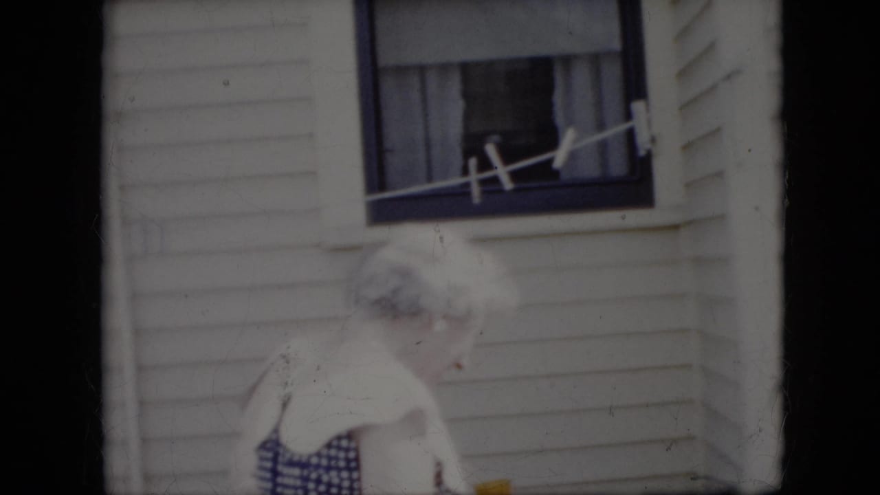 Two elderly women stand close together outside a white-sided California house in 1969, with a window, curtains, and a line visible above them.