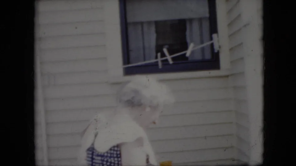 Two elderly women stand close together outside a white-sided California house in 1969, with a window, curtains, and a line visible above them.