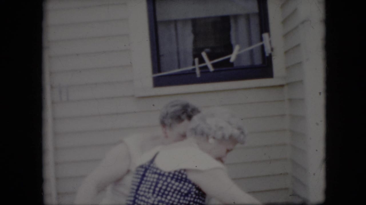 Two elderly women are seen in soft 8mm focus outside a white-sided California residence, moving near a window with a line stretched across it.
