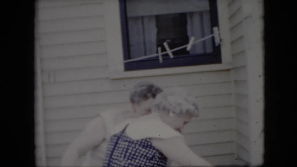 Two elderly women are seen in soft 8mm focus outside a white-sided California residence, moving near a window with a line stretched across it.