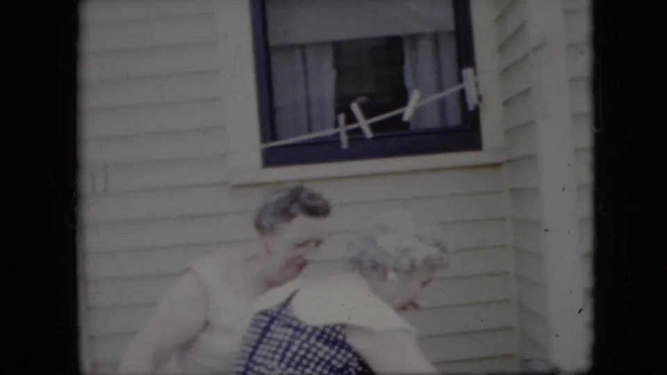Two elderly women, one in a blue checkered dress, are framed below a window on a white-sided California house in supplied 1969 home-movie footage.