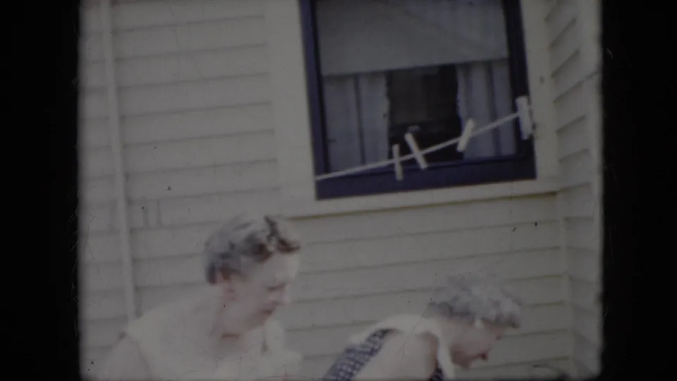 Two elderly women appear to lean or step together outside a white-sided California house, with the window and line above them visible in the 1969 home-movie frame.