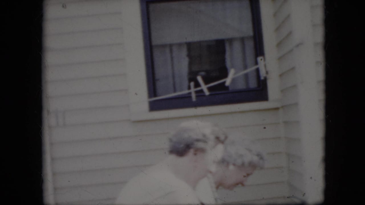 Two older women in light clothing appear beside a clothesline and curtained window outside a California house in 1969 home-movie footage.