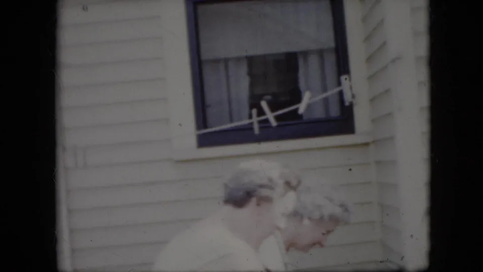 Two older women in light clothing appear beside a clothesline and curtained window outside a California house in 1969 home-movie footage.
