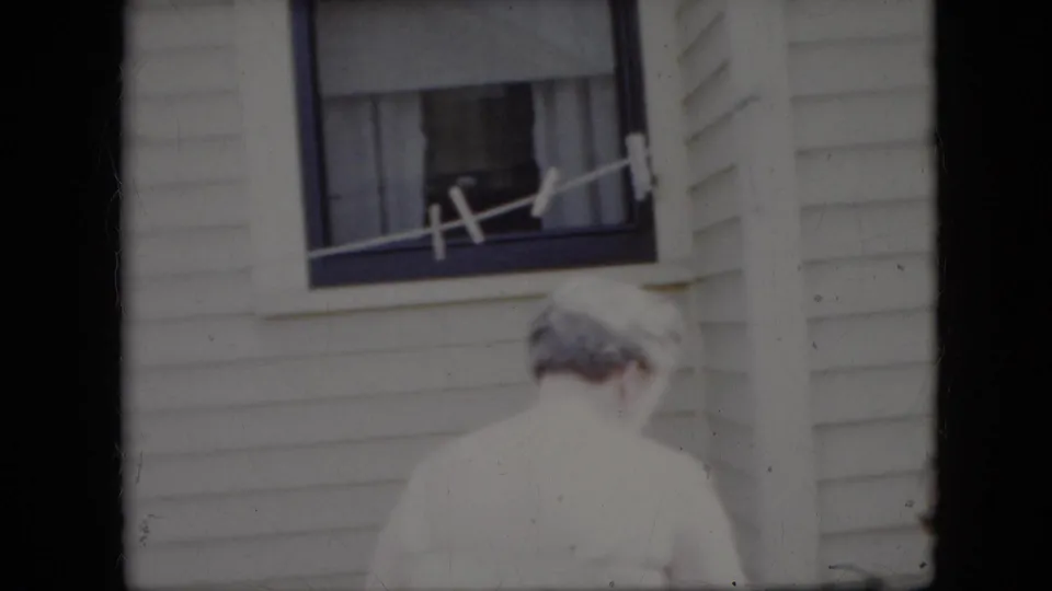 An older woman with gray hair faces the white house siding below a curtained window and clothesline in 1969 California home-movie footage.