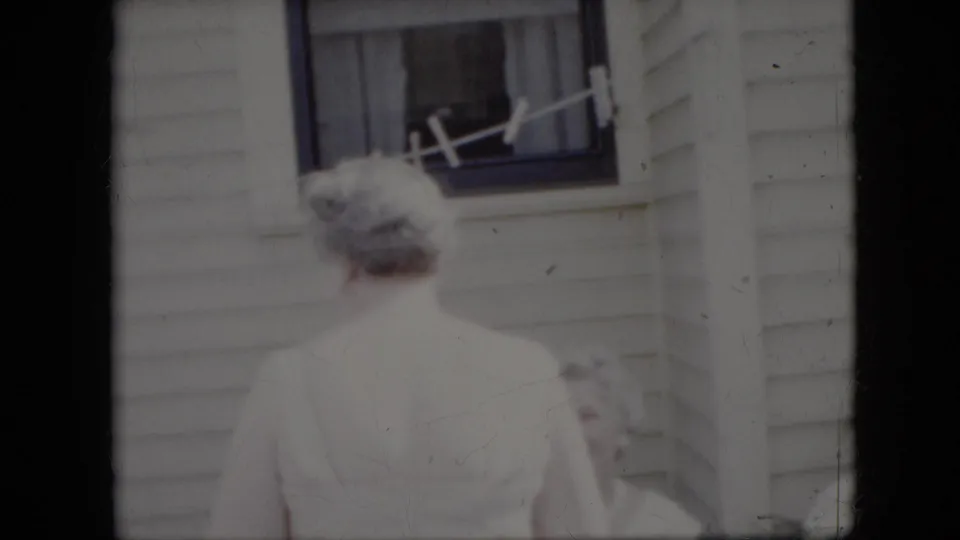 An older woman in pale clothing stands with her back to the camera near the clothesline and window of a white-sided house in California, 1969.