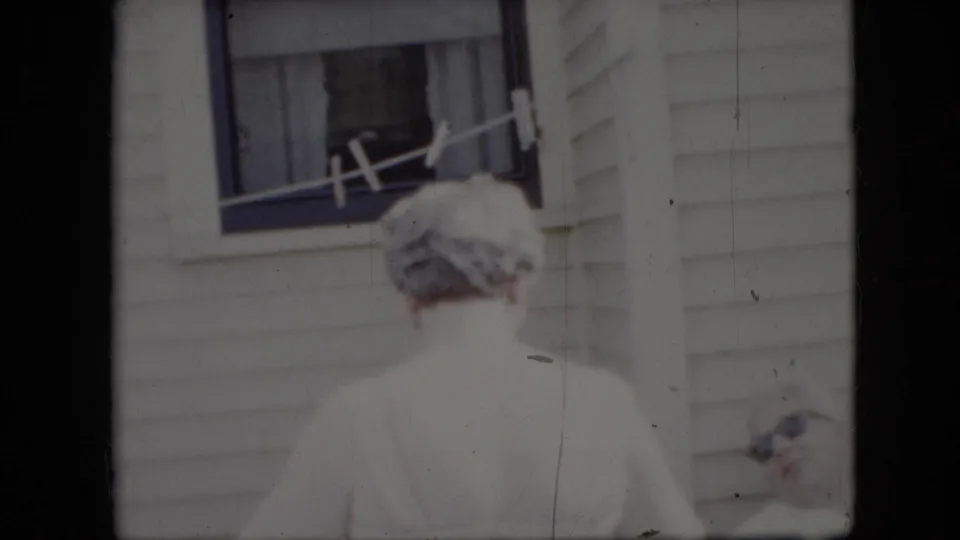 An older woman stands below a clothesline outside a white-sided California house, with another older person partly visible at the lower right edge of the 1969 home-movie frame.