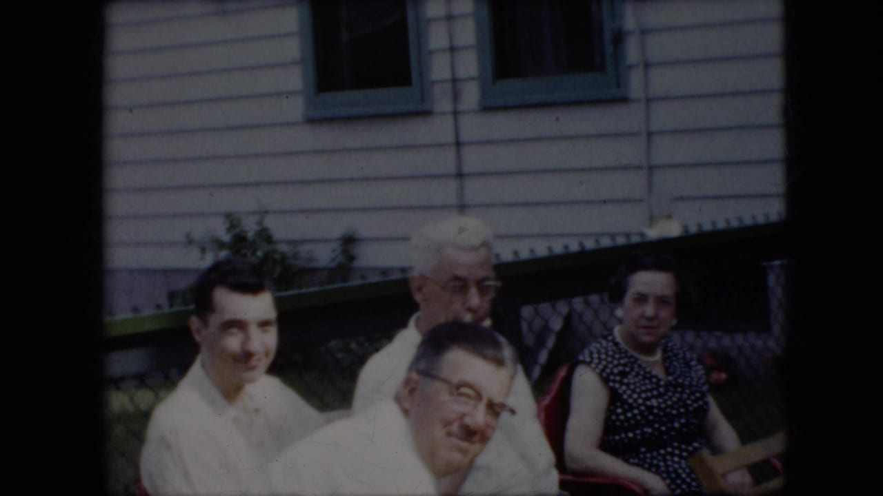 1969 California home-movie frame of four adults seated outdoors near a white-sided house with green trim, including two men in light shirts and women in patterned dresses.