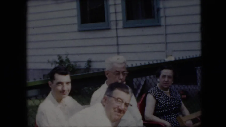 1969 California home-movie frame of four adults seated outdoors near a white-sided house with green trim, including two men in light shirts and women in patterned dresses.