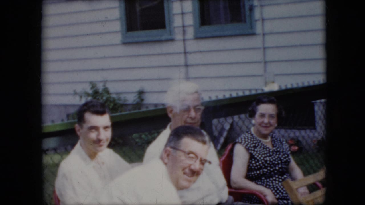 1969 California home-movie frame showing adults seated on outdoor chairs, several looking toward the camera beside house siding, windows, green trim, and a fence.