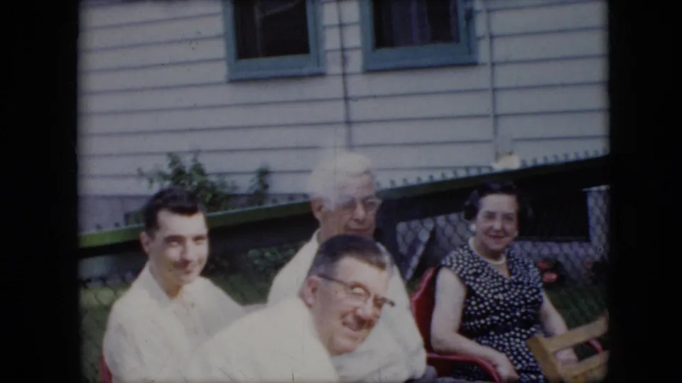 1969 California home-movie frame showing adults seated on outdoor chairs, several looking toward the camera beside house siding, windows, green trim, and a fence.