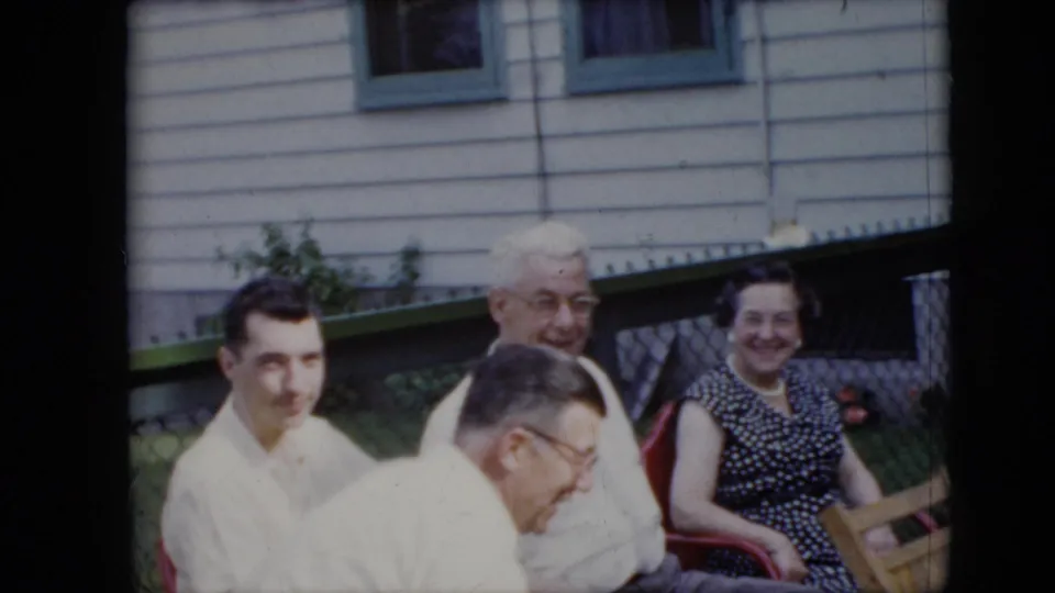 1969 California home-movie frame of adults seated together outdoors as one man bends forward, with house siding, green trim, windows, and red seating visible behind them.