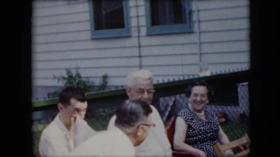 1969 California home-movie frame showing a candid outdoor family-gathering moment as seated adults shift positions near a white-sided house and chain-link fence.