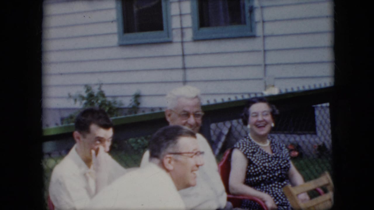Adults seated outdoors beside a white-sided house, with two men in light shirts and a woman smiling in a polka-dot dress.