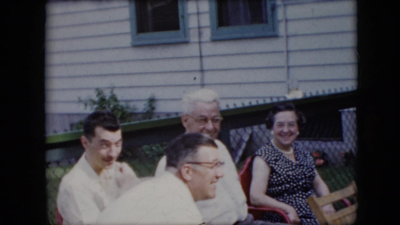 Group of adults seated in a backyard, including an older man and a smiling woman near a chain-link fence.