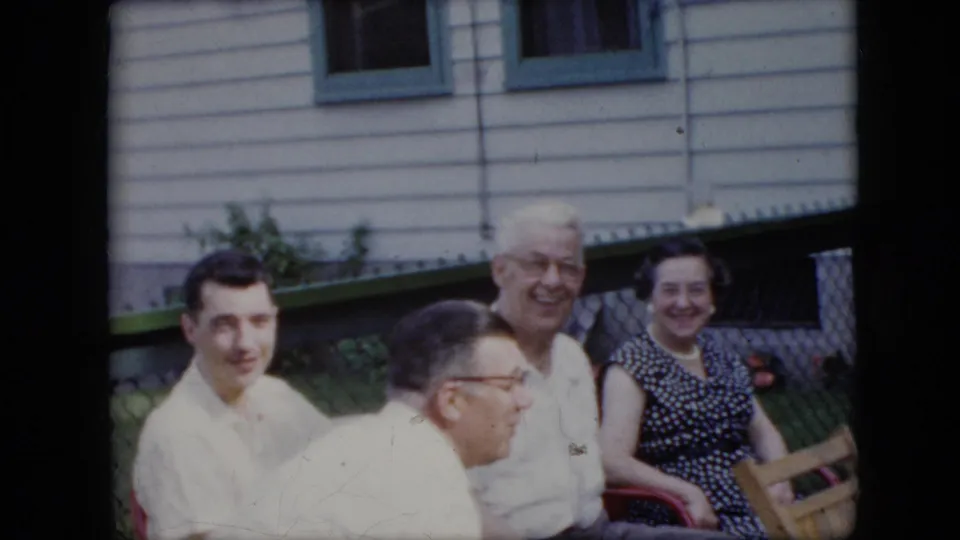 1969 California 8mm frame showing a candid backyard gathering, with adults seated close together and one man looking toward the camera.