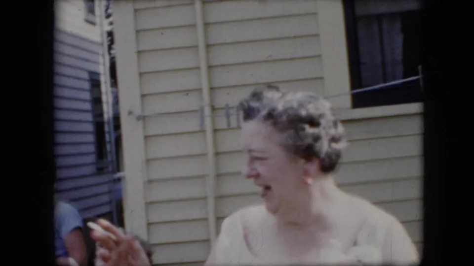 1969 California home-movie frame focusing on a woman laughing in a backyard beside a residential exterior and clothesline.