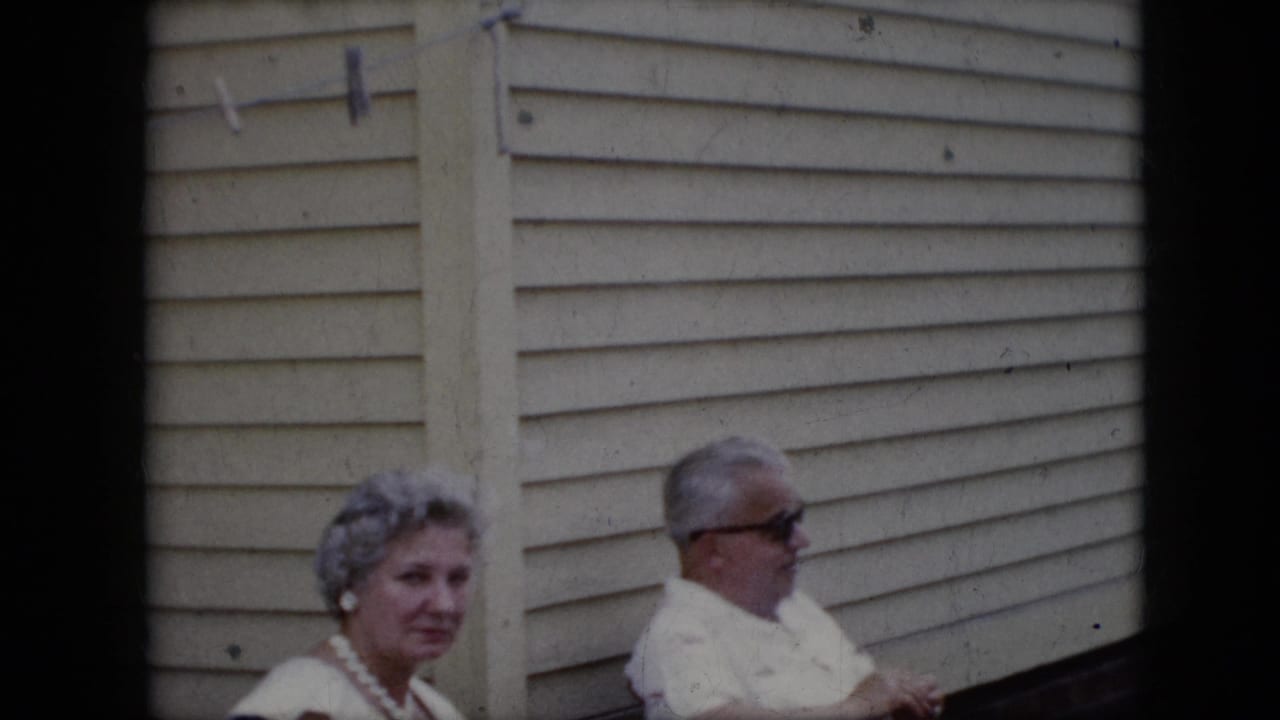 1969 California home-movie frame showing an older woman with styled gray hair and pearls seated near an older man in sunglasses outside a house with light siding.