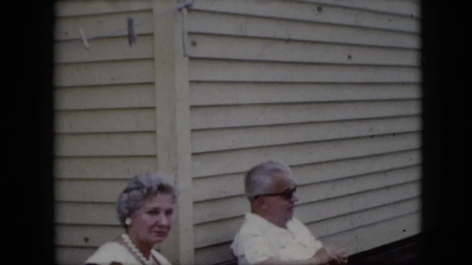 1969 California home-movie frame showing an older woman with styled gray hair and pearls seated near an older man in sunglasses outside a house with light siding.