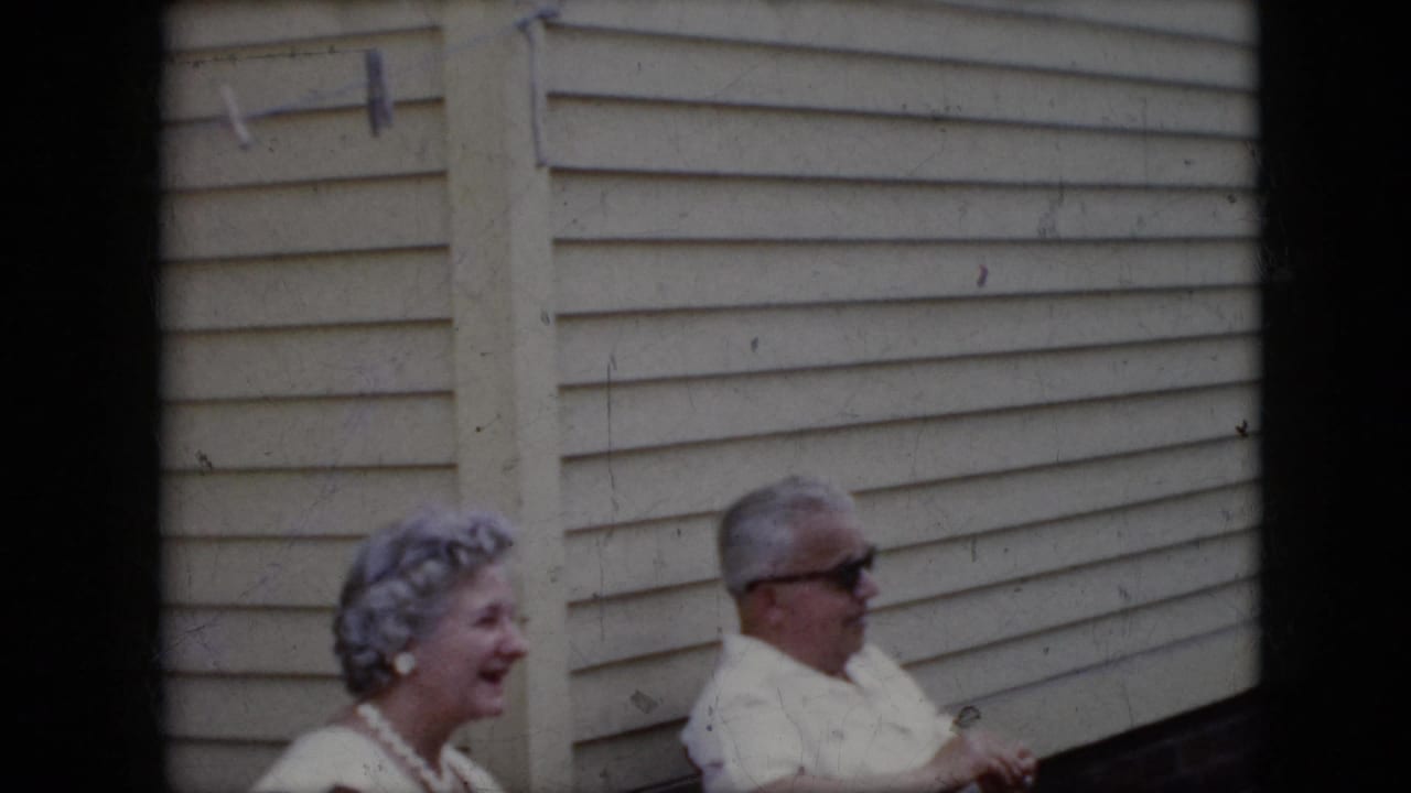 1969 California home-movie frame of an older woman smiling as she sits beside an older man in sunglasses against light exterior siding.