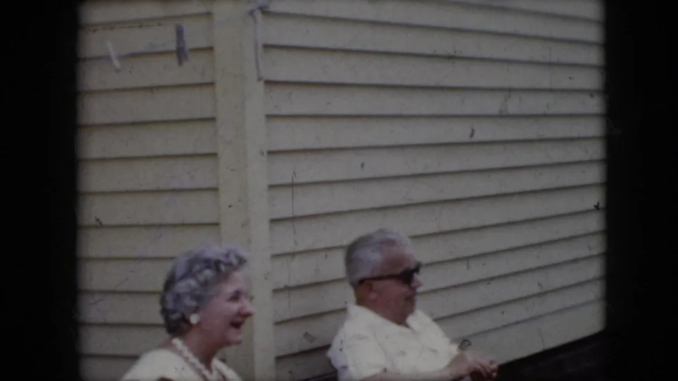 1969 California home-movie frame of an older woman smiling as she sits beside an older man in sunglasses against light exterior siding.