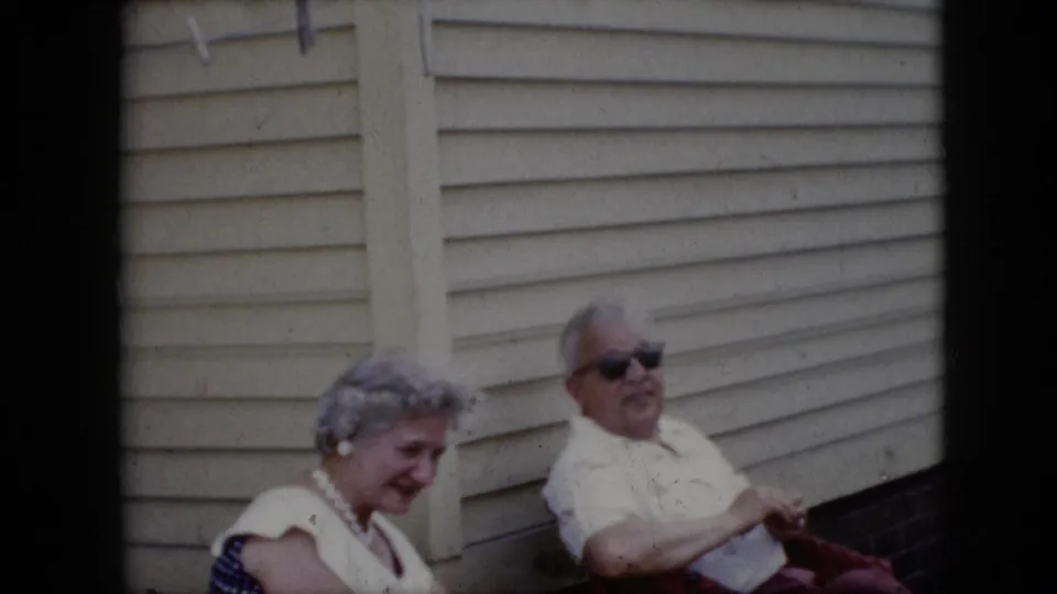 1969 California home-movie frame showing an older couple seated outdoors, with the man in dark sunglasses facing the camera and the woman looking downward near clapboard siding.