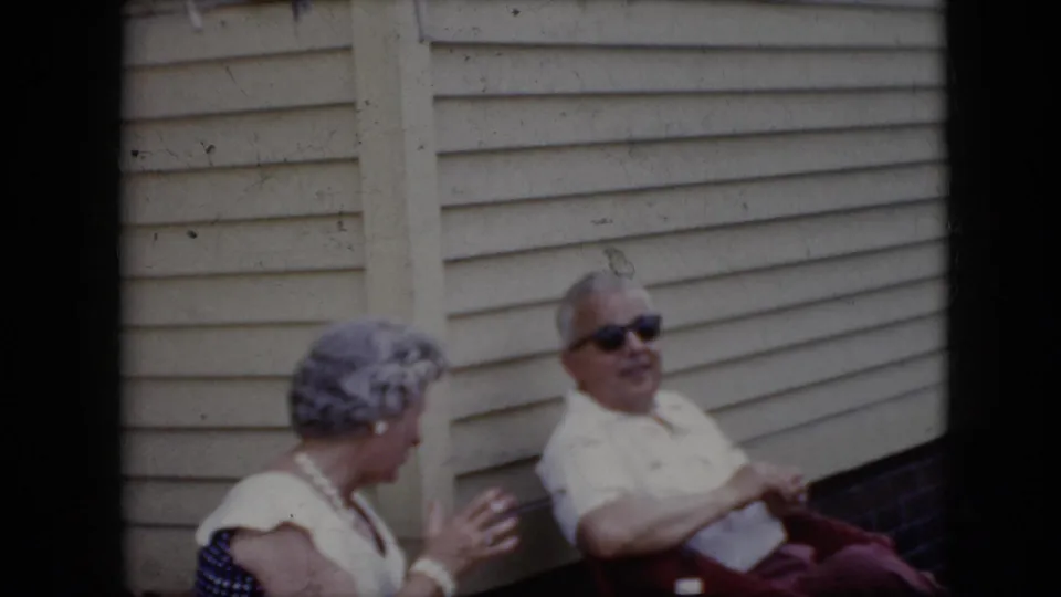 1969 California home-movie frame showing an older woman turned toward a seated man in sunglasses outside a light-sided house, suggesting casual conversation.