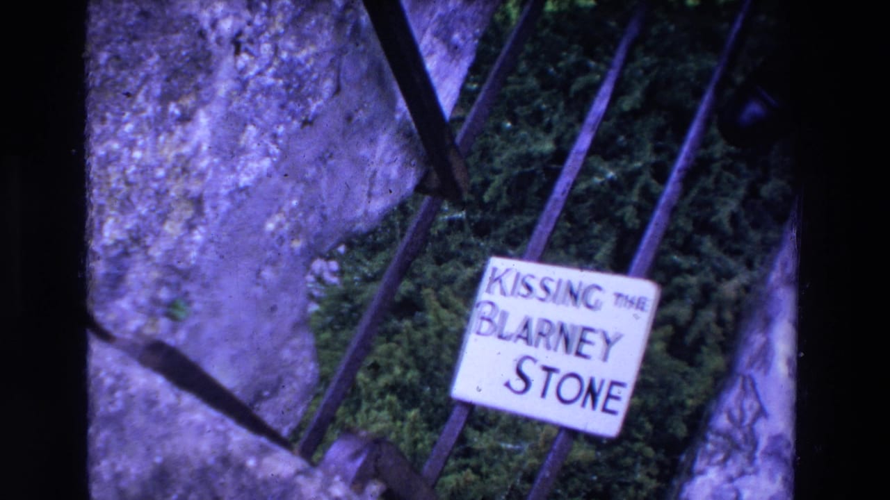 1969 Ireland 8mm frame showing a Kissing the Blarney Stone sign on a black metal railing near rough stonework and green foliage.