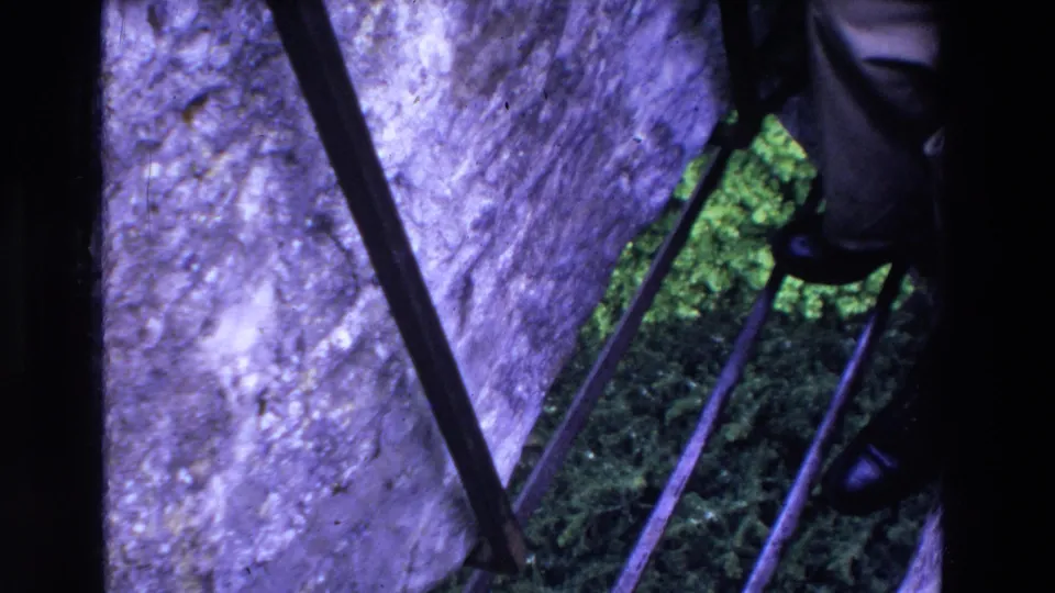 1969 Ireland 8mm frame showing the Blarney Stone railing area, rough stonework, foliage, and a partial visitor lower leg with dark shoe at the right edge.