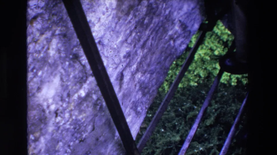 1969 Ireland 8mm frame looking down through dark metal fence bars toward purple-tinted stone and dense green moss or grass.