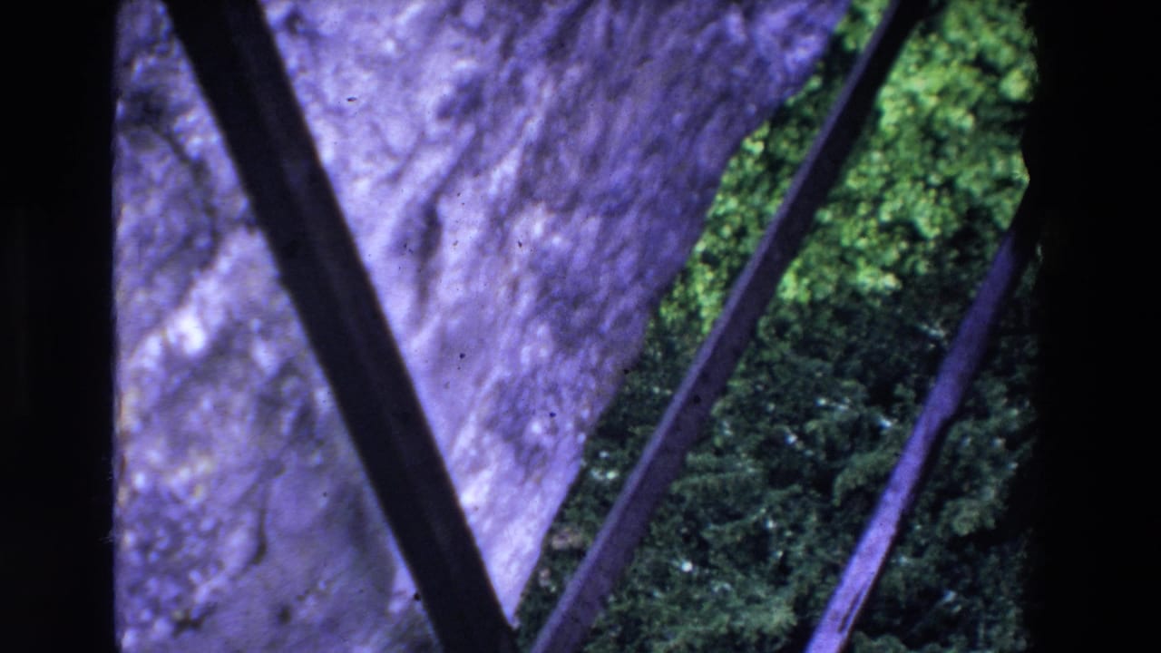 1969 Ireland 8mm frame with diagonal metal railing crossing a close view of stone, moss, and green ground below.