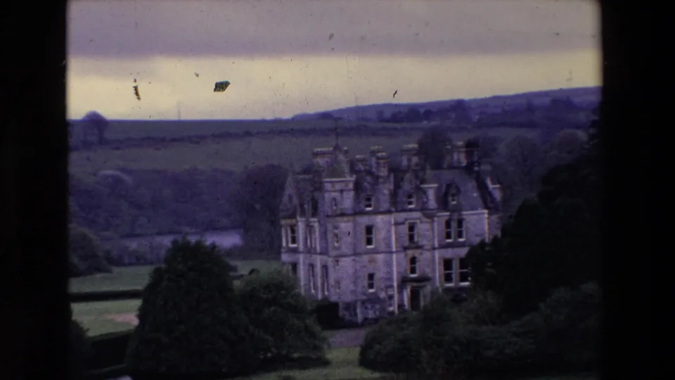 A grand stone castle appears in the Irish countryside, framed by lawns, shrubs, fields, and distant hills in 1969 8mm footage.