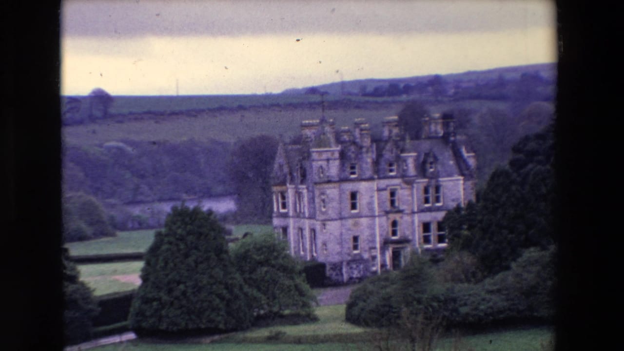 1969 8mm view in Ireland of a turreted stone country house set below rolling hills with lawns and dense trees in the foreground.