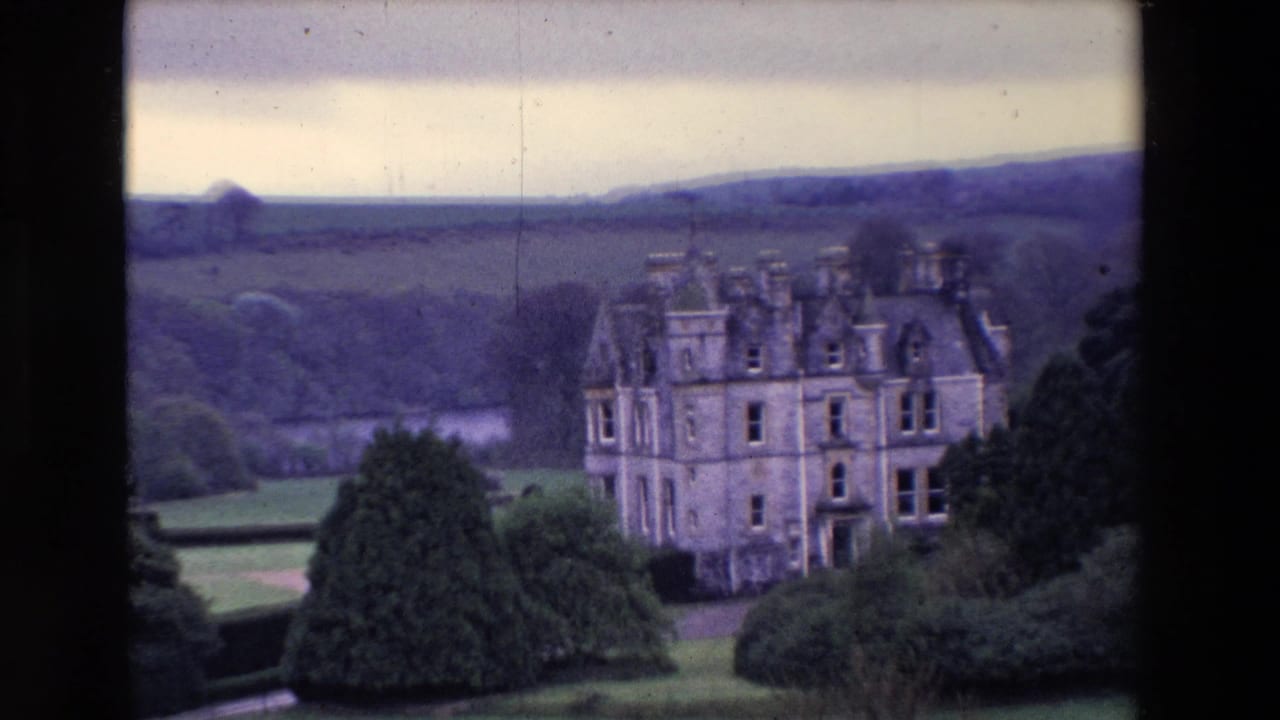Turreted stone building framed by dark trees and distant hills.