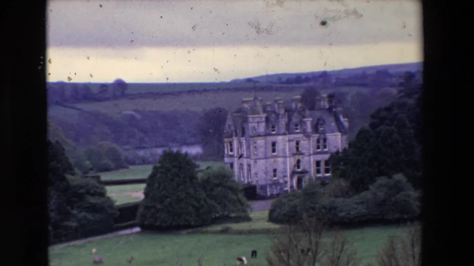 1969 Ireland home-movie frame of a castle-like country house below a hillside, with clipped lawns, shrubs, mature trees, and visible film dust across the sky.