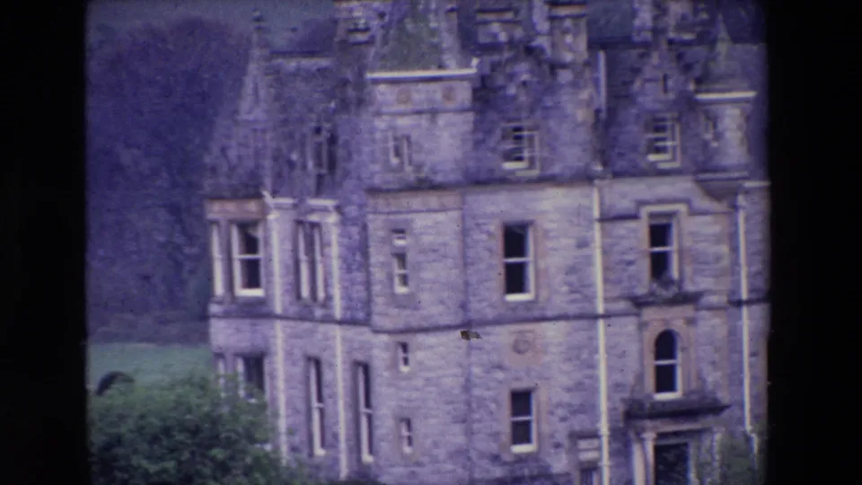 Closer 1969 Ireland home-movie view of a stone castle facade, showing a turreted corner, tall windows, dormers, roof details, and grass below.