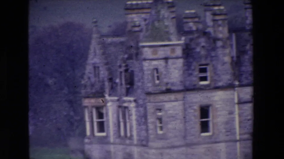 Soft-focused 1969 Ireland home-movie close view of the castle's upper stone facade, with dormer windows, turreted roofline, vertical windows, and wooded background.