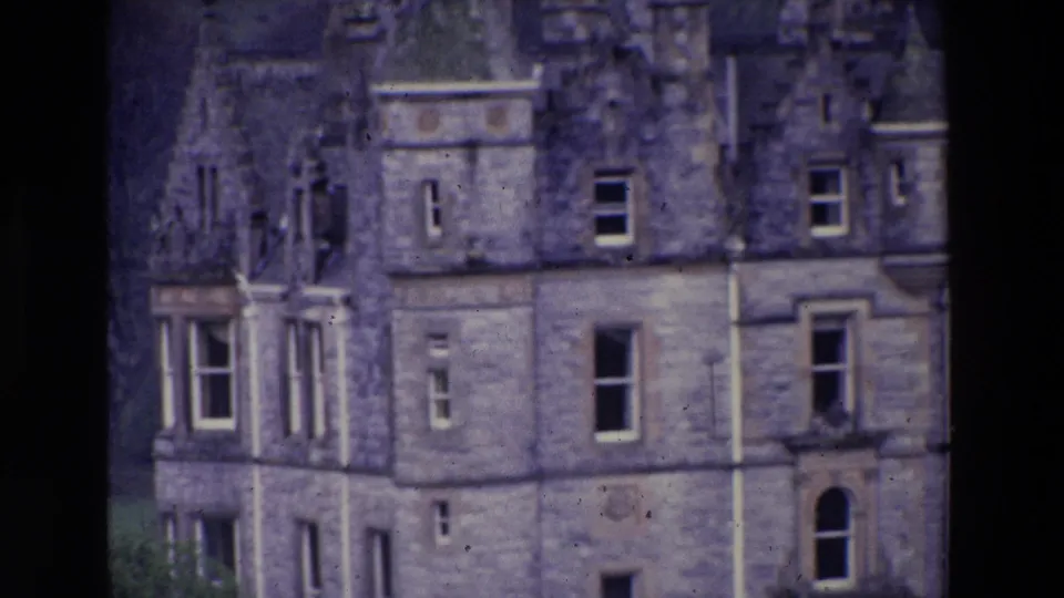 Closer 1969 Ireland home-movie view of the castle exterior, showing weathered stone walls, a turreted corner, several windows, roof dormers, and lawn at the base.