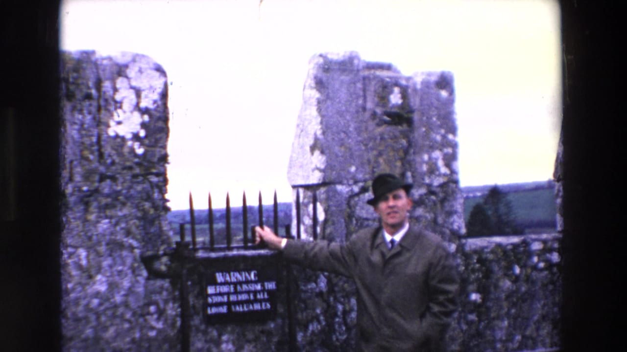 Adult man in a trench coat and dark hat stands beside a black pointed iron gate between weathered stone pillars in Ireland.