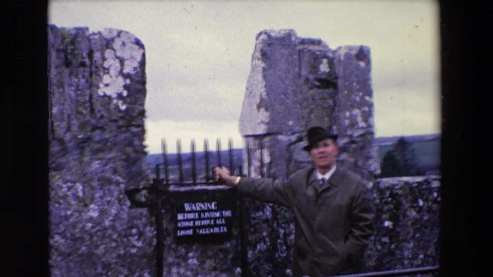 The man holds or gestures toward the black iron gate near a warning sign, with rough stone pillars and open landscape behind him.