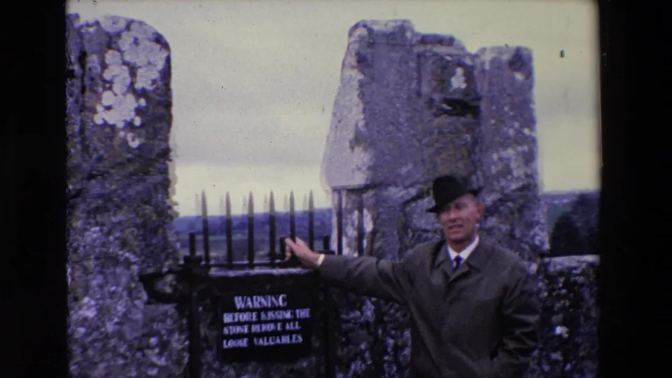 Adult man in a coat and hat stands at the stone gate with a visible warning sign, iron spikes, rough masonry, and distant countryside.
