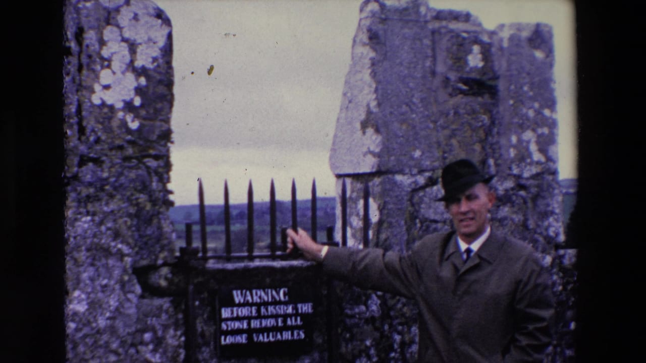 In 1969 Ireland home-movie footage, a man in a dark hat and trench coat stands beside a weathered stone wall, pointing toward a warning sign below an iron gate.
