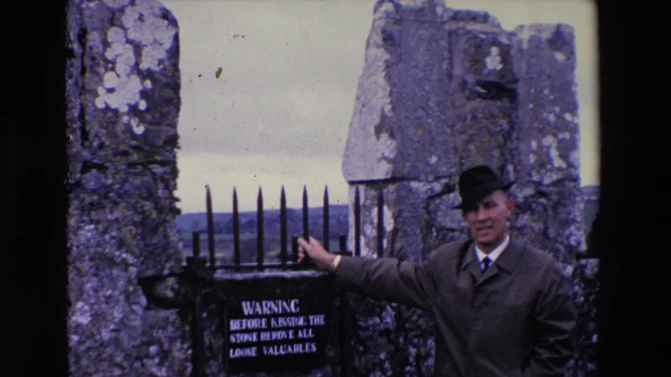 In 1969 Ireland home-movie footage, a man in a dark hat and trench coat stands beside a weathered stone wall, pointing toward a warning sign below an iron gate.