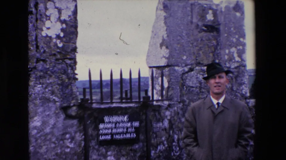In 1969 Ireland, a man in a dark coat and brimmed hat poses near weathered stone walls, a black iron gate, and a sign warning visitors before kissing the stone.