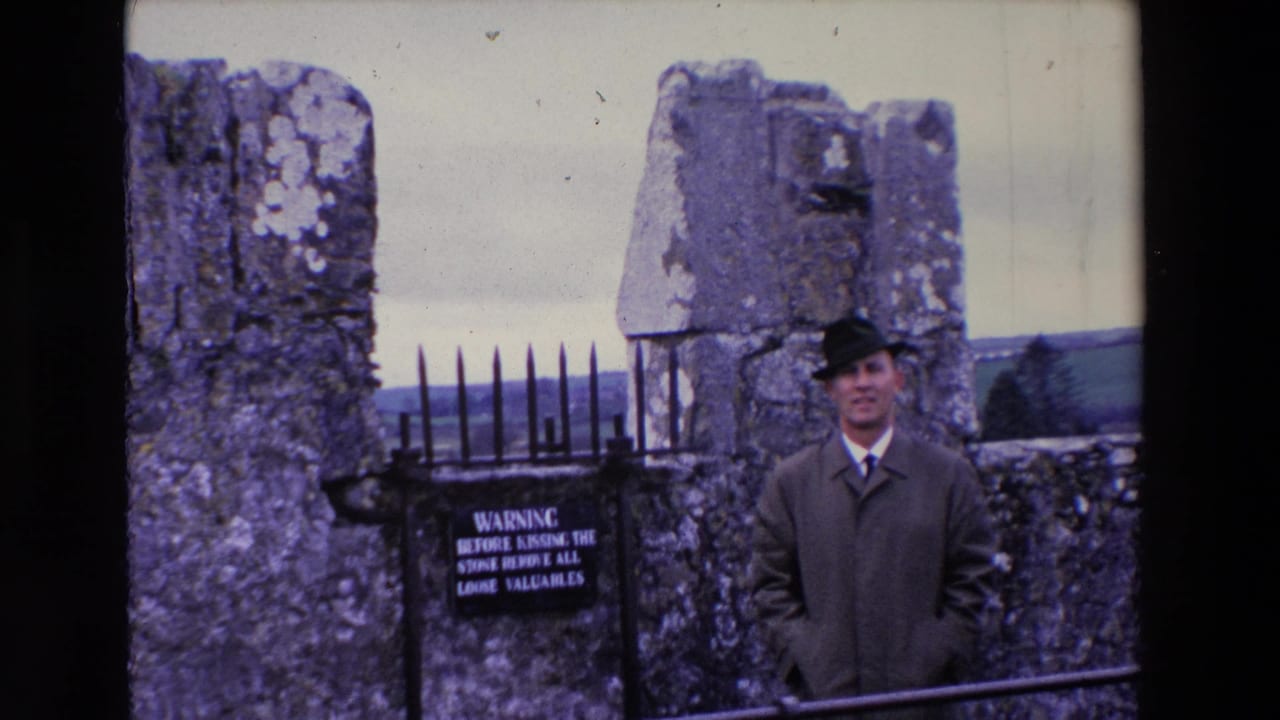 In 1969 Ireland, the man remains posed near a black iron fence and warning sign, with rough stonework and a rural horizon behind him.