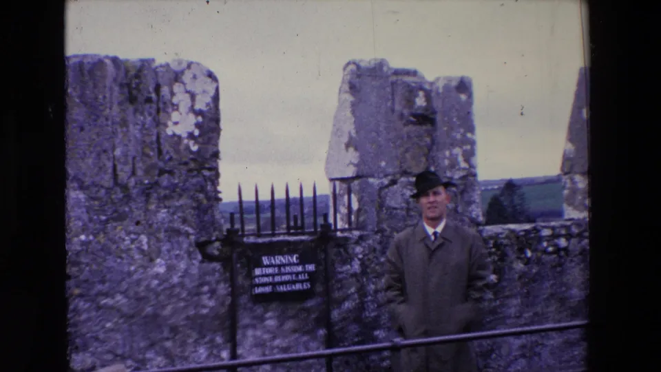 In 1969 Ireland, the camera frames the man lower beside the iron rail, with the warning sign, rough stone walls, and open countryside still visible.