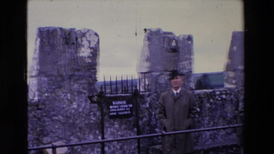 In 1969 Ireland, the man stands behind a foreground iron rail at the historic stone site, with the black warning sign and lichen-covered stonework beside him.