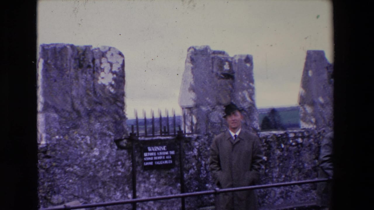 In 1969 Ireland, a man in a dark coat and hat stands near weathered Blarney Castle stonework, metal railings, a spiked gate, and a visible warning sign.