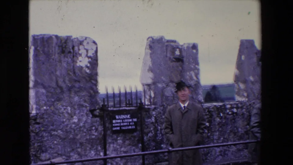 In 1969 Ireland, a man in a dark coat and hat stands near weathered Blarney Castle stonework, metal railings, a spiked gate, and a visible warning sign.