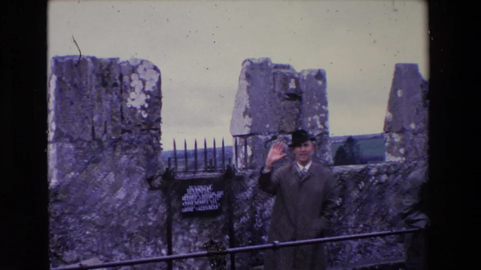 In 1969 Ireland, a man at Blarney Castle raises his hand toward the camera while standing near the stone ruins, warning sign, and metal railing.